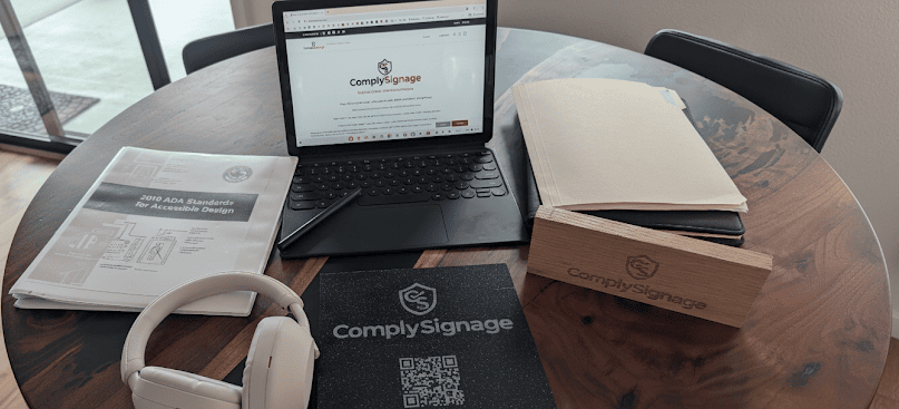 A laptop with the ComplySignage logo displayed on the screen, surrounded by documents, a wooden block with the company name, and a pair of white headphones.