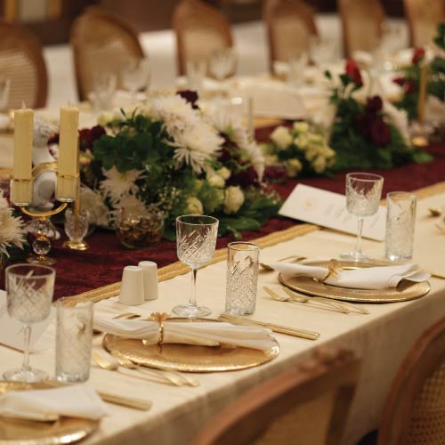Long ceremonial dining table with cream linen, burgundy runner and gold place settings Long ceremonial dining table with cream linen, burgundy runner and gold place settings