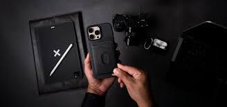 Persona sosteniendo un iPhone 15 Pro Max con funda protectora negra, acompañado de accesorios tecnológicos en un escritorio negro en Pereira, Colombia. Persona sosteniendo un iPhone 15 Pro Max con funda protectora negra, acompañado de accesorios tecnológicos en un escritorio negro en Pereira, Colombia.