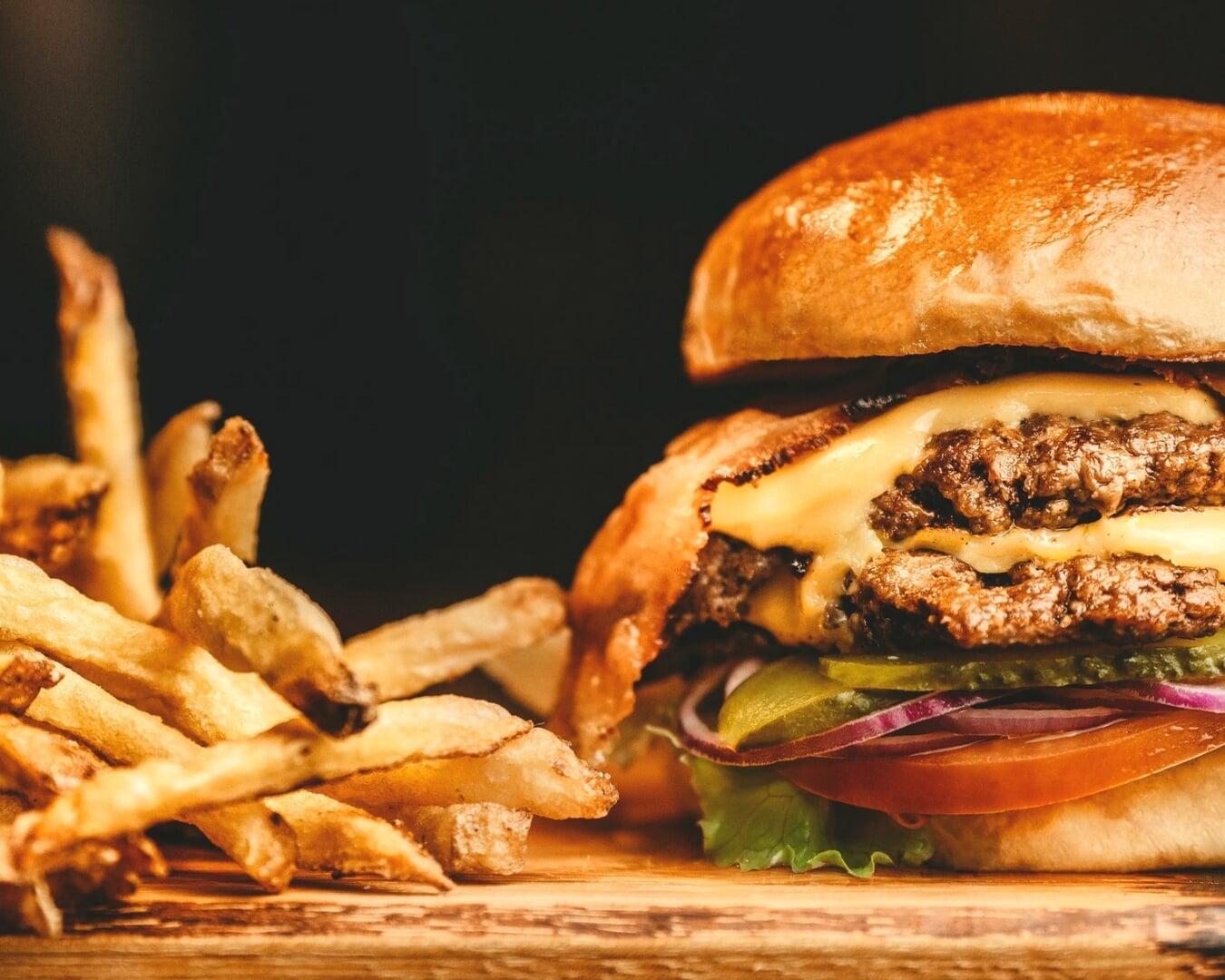 A close-up view of a double cheeseburger—two beef patties with melted cheese, lettuce, tomato, pickles, and a toasted bun—sitting on a wooden surface alongside a pile of golden, crispy French fries against a dark background. A close-up view of a double cheeseburger—two beef patties with melted cheese, lettuce, tomato, pickles, and a toasted bun—sitting on a wooden surface alongside a pile of golden, crispy French fries against a dark background.